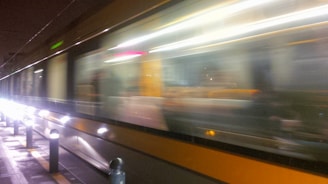 A high-speed train moves swiftly along its tracks, creating a blurred effect with bright lights reflecting off its surface. The background is dark, emphasizing the motion and speed of the train.