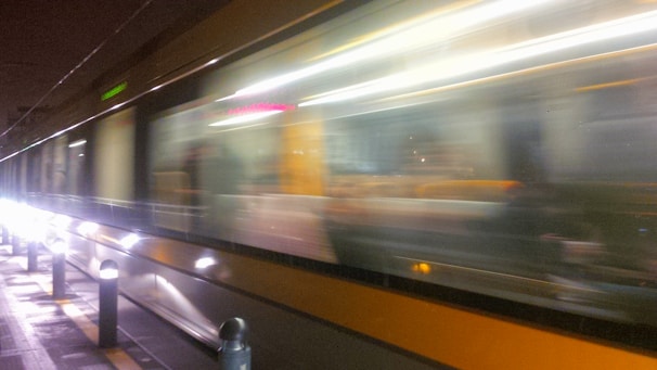 A high-speed train moves swiftly along its tracks, creating a blurred effect with bright lights reflecting off its surface. The background is dark, emphasizing the motion and speed of the train.