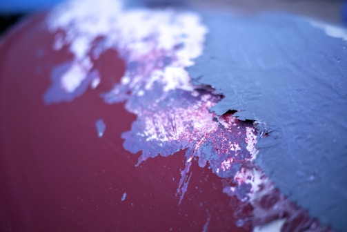 The image shows a close-up of a surface with peeling paint. The paint appears worn, with layers of different colors visible, such as blue and maroon. Some areas show signs of rust or wear.