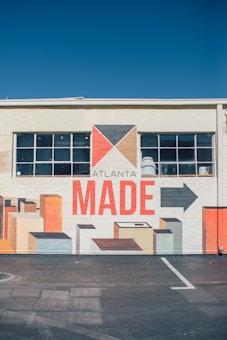 A mural painted on the side of a building, featuring geometric shapes in earth tones with a prominent text reading 'ATLANTA MADE'. The mural includes a stylized arrow and rectangular forms that resemble blocks or buildings. The building has a row of windows above the mural.