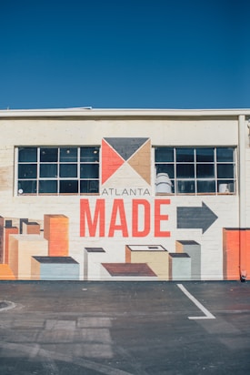 A mural painted on the side of a building, featuring geometric shapes in earth tones with a prominent text reading 'ATLANTA MADE'. The mural includes a stylized arrow and rectangular forms that resemble blocks or buildings. The building has a row of windows above the mural.