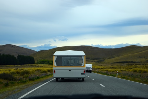 A caravan is traveling on a rural highway flanked by rolling hills and fields. In the distance, a line of trees under a cloudy sky enhances the scene’s tranquility. The road is clear except for another vehicle following behind.