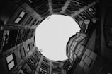 A circular view of a building's interior courtyard seen from below, featuring tall walls with numerous windows and balconies. The architectural lines form a dramatic upward perspective culminating in a bright, circular opening at the top.