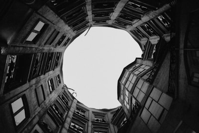 A circular view of a building's interior courtyard seen from below, featuring tall walls with numerous windows and balconies. The architectural lines form a dramatic upward perspective culminating in a bright, circular opening at the top.
