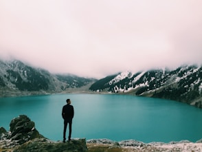 man standing on mountain looking at lake under white clouds at daytime