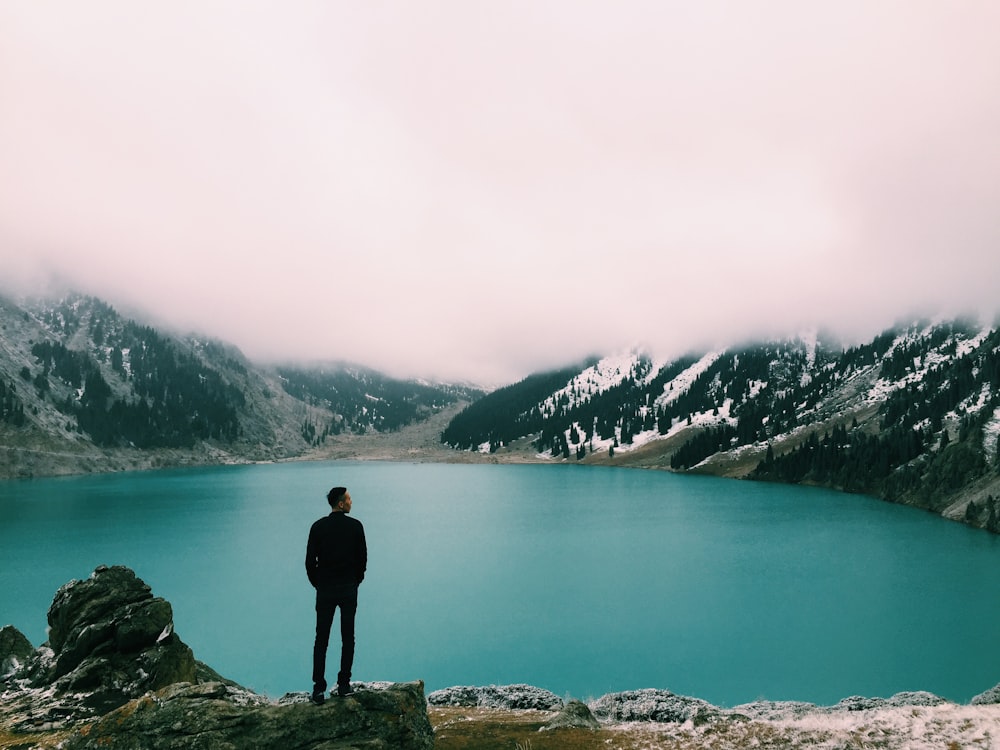 homem de pé na montanha olhando para o lago sob nuvens brancas durante o dia