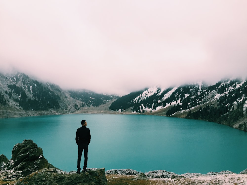 man standing on mountain looking at lake under white clouds at daytime