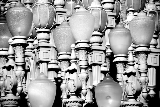 A multitude of ornate and vintage-style street lamps are clustered together, displaying intricate designs and varied shapes. The lamps are made of metal and glass and are arranged in a dense, overlapping manner, creating an artistic and textured visual effect. The monochrome color scheme adds a classic and timeless feel to the composition.