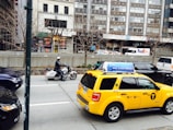 A busy urban street scene featuring a police officer on a motorcycle and a yellow taxi with advertisements on the roof. In the background, there are tall buildings, a FedEx truck, and construction with scaffolding. Leafless trees line the sidewalk, indicating a colder season.