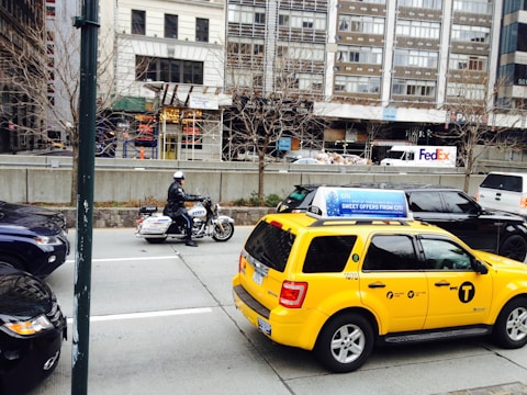 A busy urban street scene featuring a police officer on a motorcycle and a yellow taxi with advertisements on the roof. In the background, there are tall buildings, a FedEx truck, and construction with scaffolding. Leafless trees line the sidewalk, indicating a colder season.