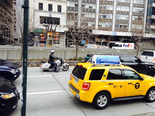 A busy urban street scene featuring a police officer on a motorcycle and a yellow taxi with advertisements on the roof. In the background, there are tall buildings, a FedEx truck, and construction with scaffolding. Leafless trees line the sidewalk, indicating a colder season.