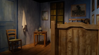 A room with blue-painted walls features rustic wooden furniture, including a chair, a small table, and a wooden panel. A white cloth hangs on the wall, and various framed artworks and a pitcher with a bowl sit on the table. The room is softly lit, with a warm, cozy ambiance.