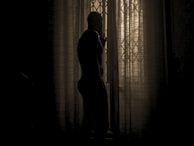 Soft-focus image of a woman standing by a window, her silhouette subtly outlined by soft daylight.