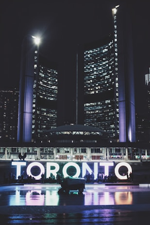 Tall, illuminated skyscrapers tower over a public square at night, with the word 'TORONTO' brightly lit in various colors at the forefront. Reflections of the lights shimmer on a wet surface below.