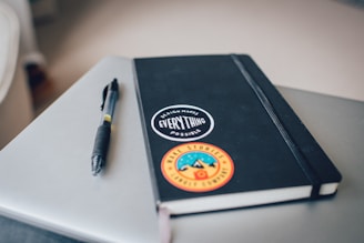 A notebook with a black cover is adorned with two stickers resting on a closed laptop. One sticker reads 'Design makes everything possible' and the other features mountains with the words 'Make stories, leave a trail'. A pen is placed beside the notebook.