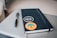 A notebook with a black cover is adorned with two stickers resting on a closed laptop. One sticker reads 'Design makes everything possible' and the other features mountains with the words 'Make stories, leave a trail'. A pen is placed beside the notebook.