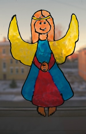 A hand-drawn image of a smiling angel with yellow wings and a golden halo, wearing a blue and red gown. The figure is set against a blurred background of buildings and sky.