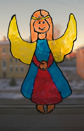 A hand-drawn image of a smiling angel with yellow wings and a golden halo, wearing a blue and red gown. The figure is set against a blurred background of buildings and sky.