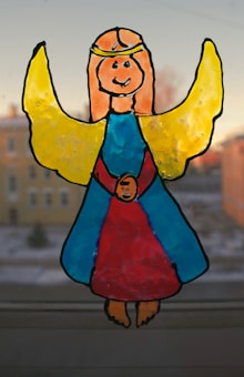 A hand-drawn image of a smiling angel with yellow wings and a golden halo, wearing a blue and red gown. The figure is set against a blurred background of buildings and sky.
