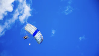 A tandem skydiver and instructor soaring above a sea of fluffy white clouds under a bright blue sky.