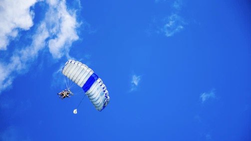 A tandem skydiver and instructor soaring above a sea of fluffy white clouds under a bright blue sky.