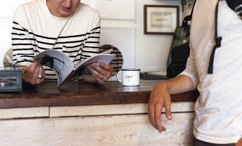 A person wearing a striped long-sleeve shirt is reading a magazine while leaning on a wooden counter. Another person, partially visible, stands nearby wearing a light-colored top. A white mug with the word 'SCOUT' printed on it is placed on the counter. The background includes framed artwork and a shelf.