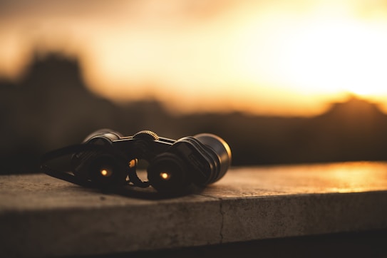 Binoculars rest on a stone surface against the backdrop of a warm, golden sunset. The focus is on the binoculars, which are softly illuminated by the ambient light, creating a serene and contemplative scene.