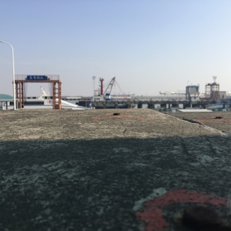 A view of an industrial port with structures such as cranes and mooring bollards. The foreground consists of a weathered concrete surface, while the background shows various maritime equipment and buildings against a clear sky.