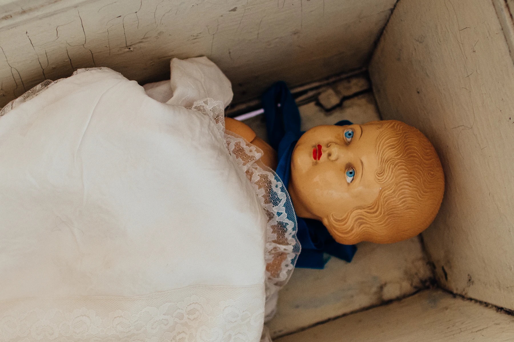An old composition doll in a white lace gown lying in a painted wooden box