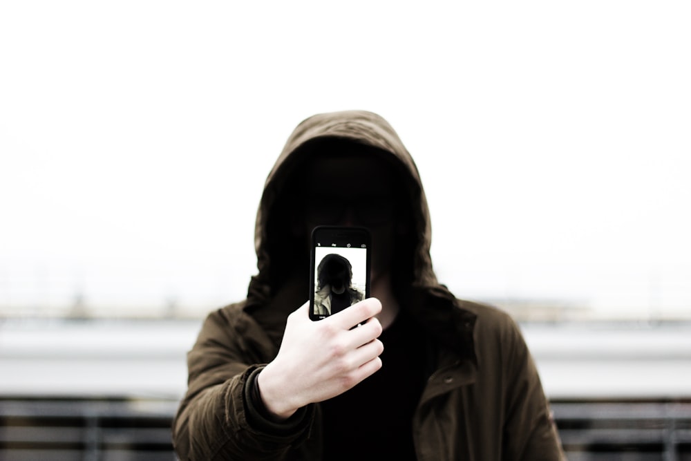homem vestindo jaqueta preta com capuz e segurando smartphone branco tirando selfie em close-up