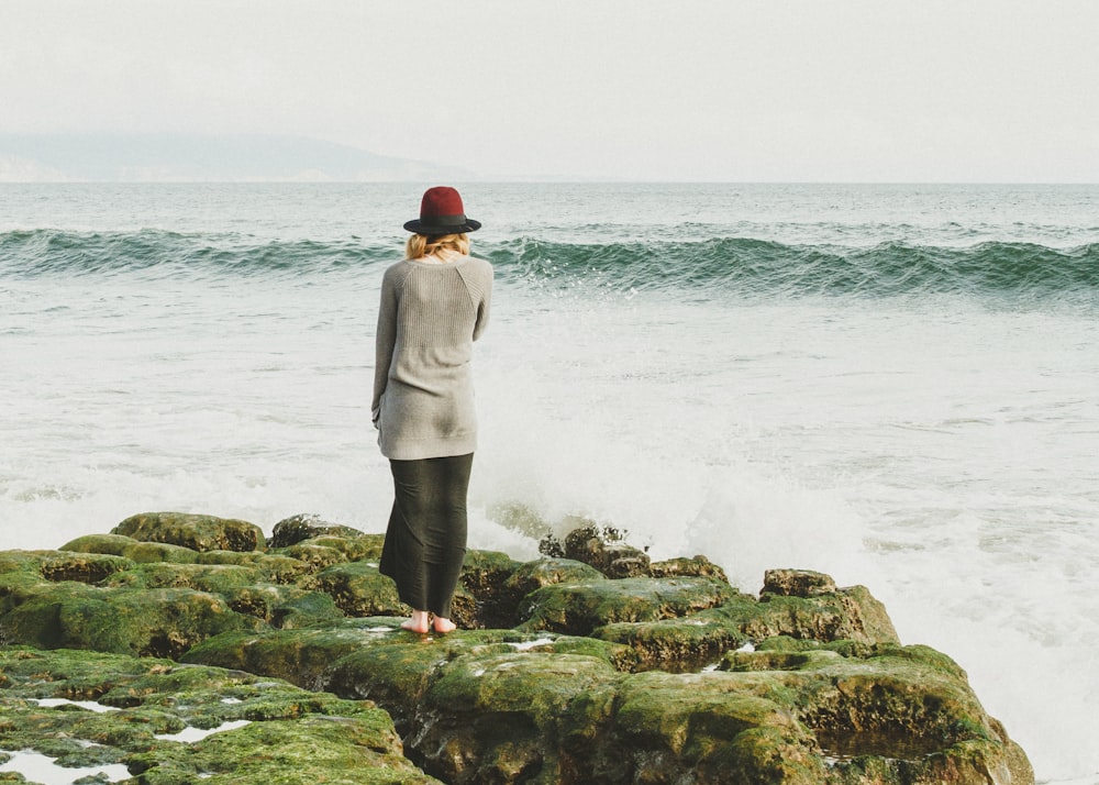 woman standing on green rocks near body of water with waves