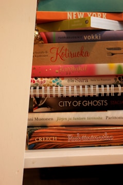 A stack of books with various colorful covers is visible on a shelf. The titles are in different languages, featuring both hardcover and spiral-bound editions. The books are arranged vertically and appear to be cookbooks, novels, and possibly travel guides.