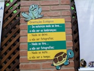 A brick wall with a wooden lattice features a colorful sign. The sign, themed with a turtle and a frog, contains ecological guidance in Portuguese text. The messages emphasize respect for nature, advising to leave no trace and to refrain from harming the environment.