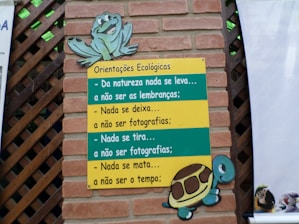 A brick wall with a wooden lattice features a colorful sign. The sign, themed with a turtle and a frog, contains ecological guidance in Portuguese text. The messages emphasize respect for nature, advising to leave no trace and to refrain from harming the environment.