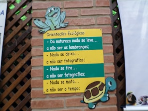 A brick wall with a wooden lattice features a colorful sign. The sign, themed with a turtle and a frog, contains ecological guidance in Portuguese text. The messages emphasize respect for nature, advising to leave no trace and to refrain from harming the environment.