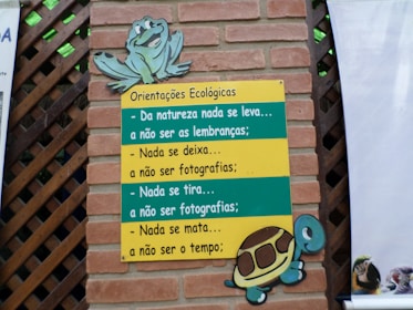 A brick wall with a wooden lattice features a colorful sign. The sign, themed with a turtle and a frog, contains ecological guidance in Portuguese text. The messages emphasize respect for nature, advising to leave no trace and to refrain from harming the environment.