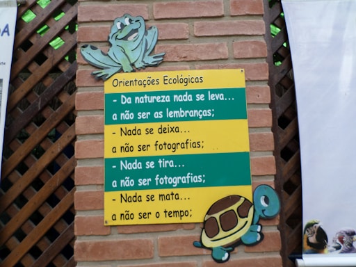 A brick wall with a wooden lattice features a colorful sign. The sign, themed with a turtle and a frog, contains ecological guidance in Portuguese text. The messages emphasize respect for nature, advising to leave no trace and to refrain from harming the environment.