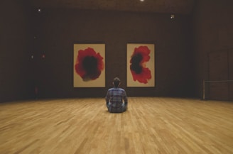 A person sits on a wooden floor in a dimly lit room, facing two large abstract paintings that feature red and black color schemes. The paintings are symmetrically hung on a textured, dark wall. The person is wearing a plaid shirt and is seated in a contemplative posture.