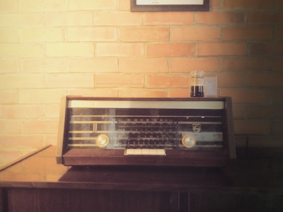 A vintage radio sits on a wooden surface against a brick wall. The radio has a dark brown frame with two gold dials and a glass display. Above the radio hangs a framed item, partially visible. A small glass jar with a dark liquid is placed beside the radio.