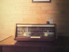 A vintage radio sits on a wooden surface against a brick wall. The radio has a dark brown frame with two gold dials and a glass display. Above the radio hangs a framed item, partially visible. A small glass jar with a dark liquid is placed beside the radio.