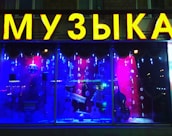 The image features a shop window display with a large, bright yellow sign displaying a word in Cyrillic letters. Inside the window, there are musical instruments including a keyboard, guitar, and double bass, set against a vibrant backdrop of red and blue lighting. Decorative snowflakes and strings of lights add a festive atmosphere.