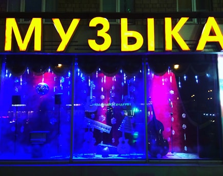 The image features a shop window display with a large, bright yellow sign displaying a word in Cyrillic letters. Inside the window, there are musical instruments including a keyboard, guitar, and double bass, set against a vibrant backdrop of red and blue lighting. Decorative snowflakes and strings of lights add a festive atmosphere.