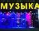 The image features a shop window display with a large, bright yellow sign displaying a word in Cyrillic letters. Inside the window, there are musical instruments including a keyboard, guitar, and double bass, set against a vibrant backdrop of red and blue lighting. Decorative snowflakes and strings of lights add a festive atmosphere.