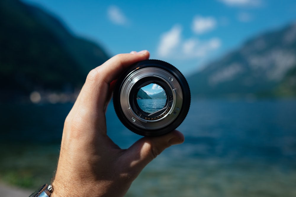 A person's hand holding a camera lens over a mountain lake