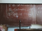 A large, worn out chalkboard filled with complex equations written in white chalk. The equations involve mathematical and physics notations, covering most of the board. The board has a reddish-brown hue with visible wear and scratches.