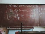A large, worn out chalkboard filled with complex equations written in white chalk. The equations involve mathematical and physics notations, covering most of the board. The board has a reddish-brown hue with visible wear and scratches.
