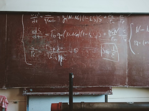 A large, worn out chalkboard filled with complex equations written in white chalk. The equations involve mathematical and physics notations, covering most of the board. The board has a reddish-brown hue with visible wear and scratches.