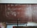 A large, worn out chalkboard filled with complex equations written in white chalk. The equations involve mathematical and physics notations, covering most of the board. The board has a reddish-brown hue with visible wear and scratches.