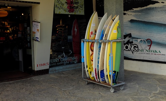 A collection of surfboards is neatly arranged in a rack outside a surf shop. The shop has a festive holiday greeting sign and posters advertising surfing events. The entrance to the shop is visible, showing a warmly lit interior with various surfing merchandise displayed. A poster on the wall features a wave with dates and text relating to a surfing competition.