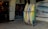 A collection of surfboards is neatly arranged in a rack outside a surf shop. The shop has a festive holiday greeting sign and posters advertising surfing events. The entrance to the shop is visible, showing a warmly lit interior with various surfing merchandise displayed. A poster on the wall features a wave with dates and text relating to a surfing competition.